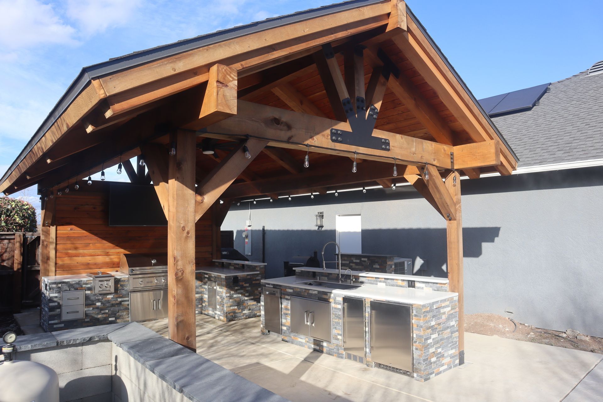 Wooden outdoor kitchen with stainless steel appliances, beneath a wood-framed roof.