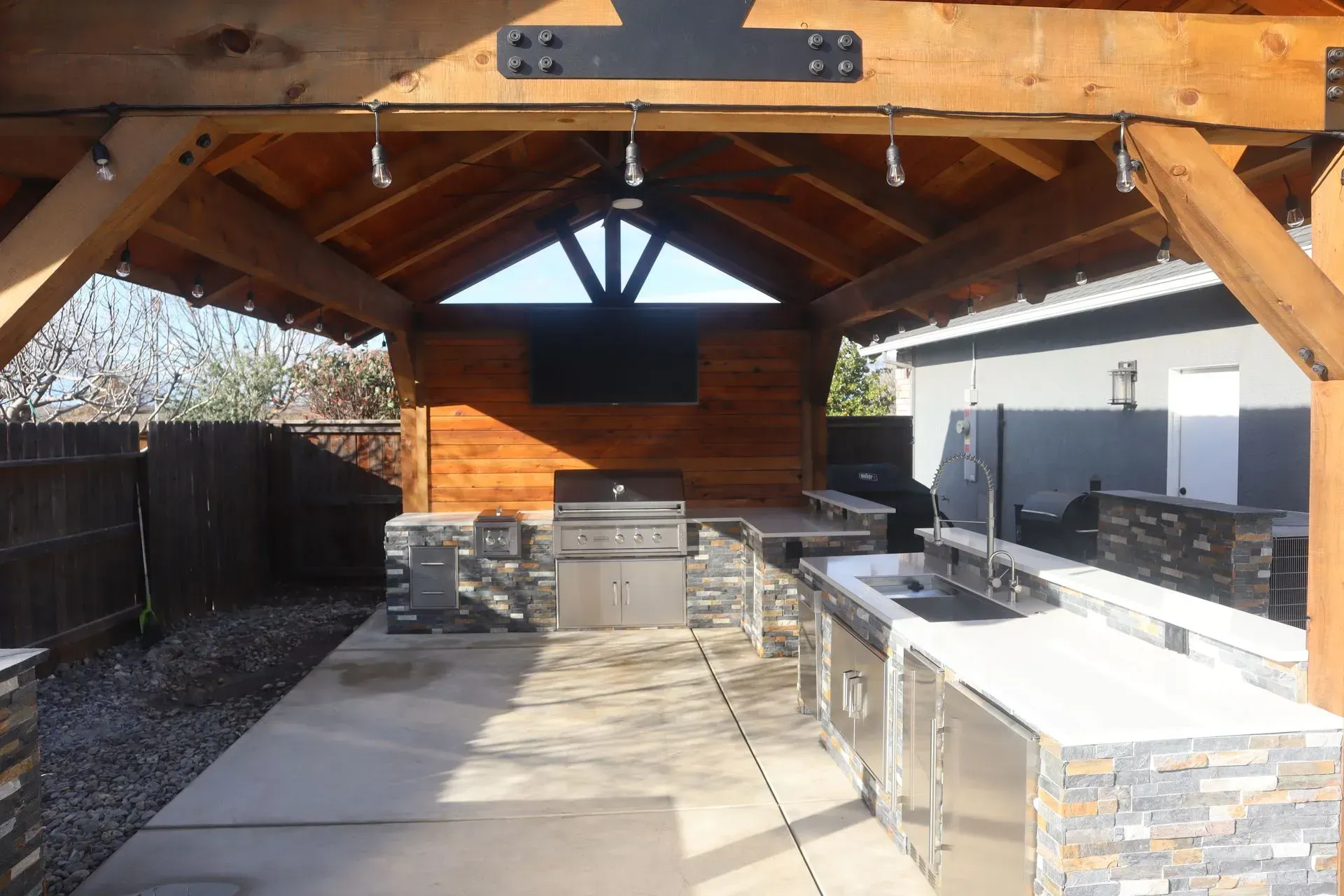 Outdoor kitchen with wooden pergola, built-in grill, countertops, and television.