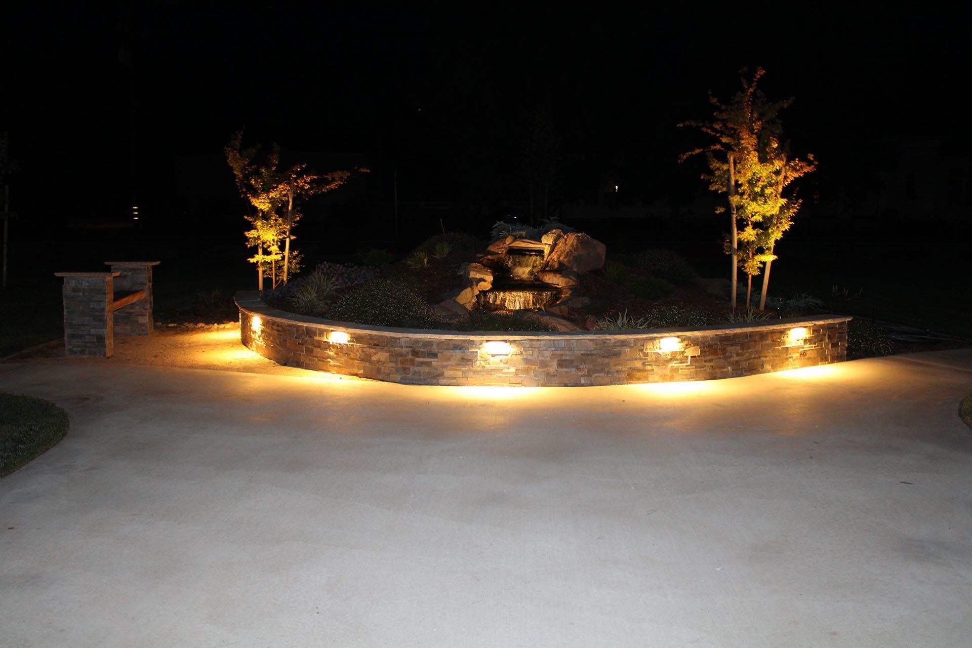 Nighttime view of a stone wall with built-in lights, surrounding a small waterfall and flanked by trees.