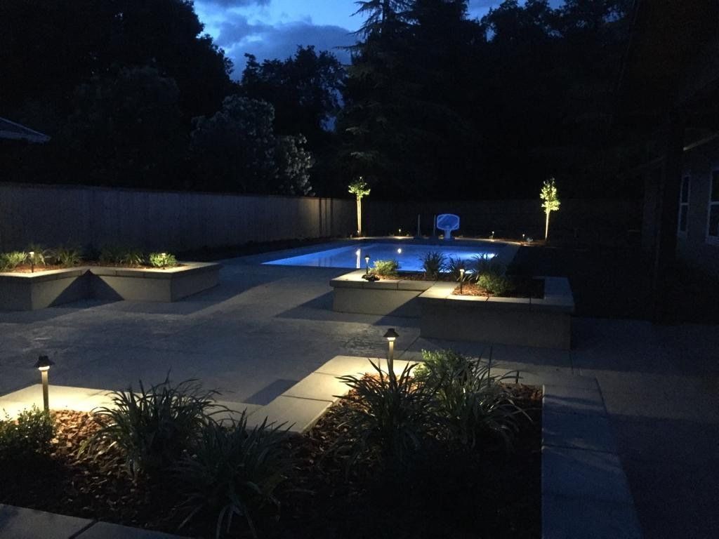 Nighttime view of a backyard pool with landscape lighting illuminating the pool, plants, and concrete patio.
