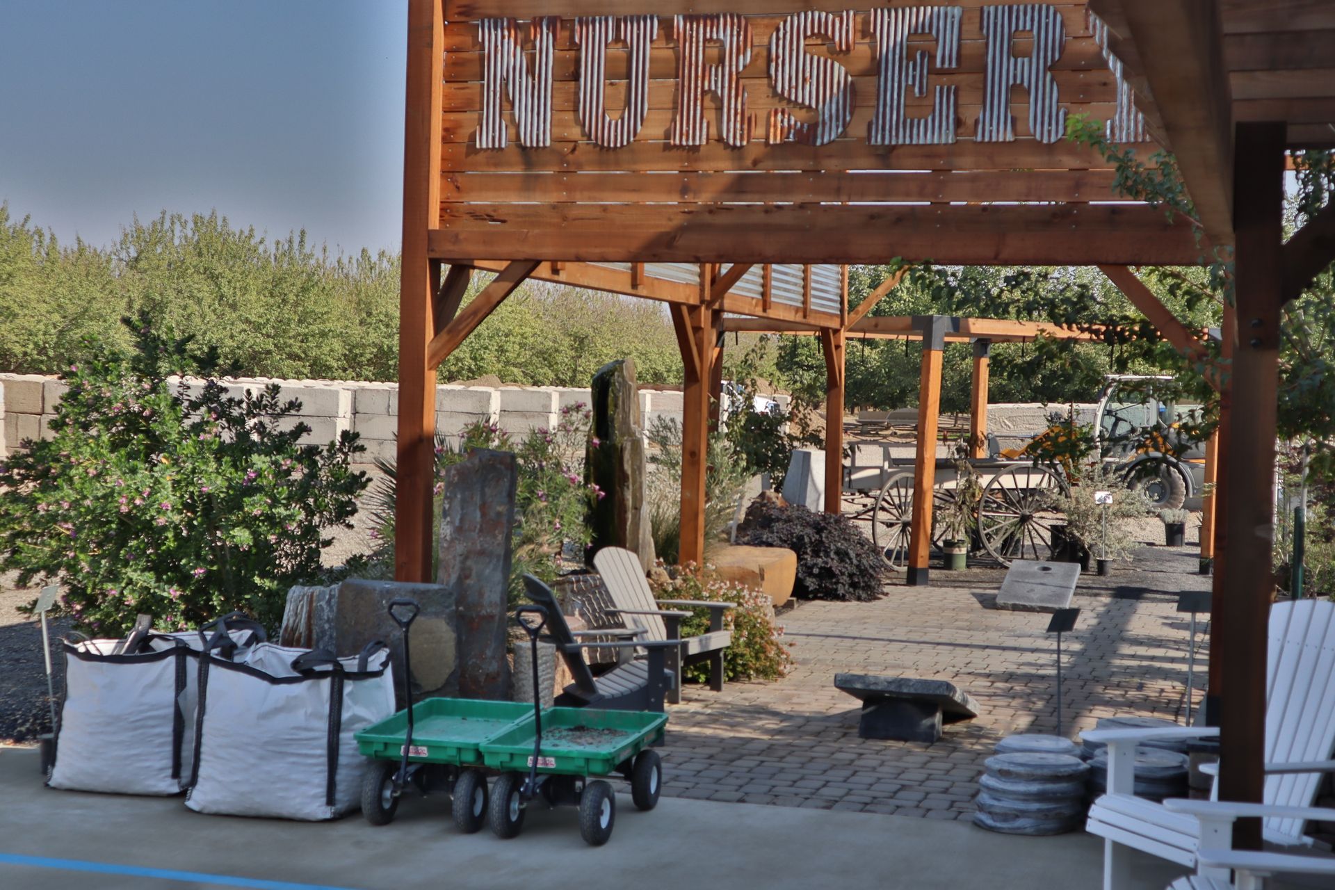 Nursery entrance with wooden sign and arbor. Two white bags, green wagon, and benches are in the foreground.
