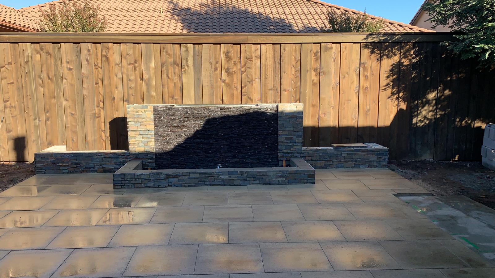 Water feature with a dark stone face on a paved patio, set against a wooden fence.