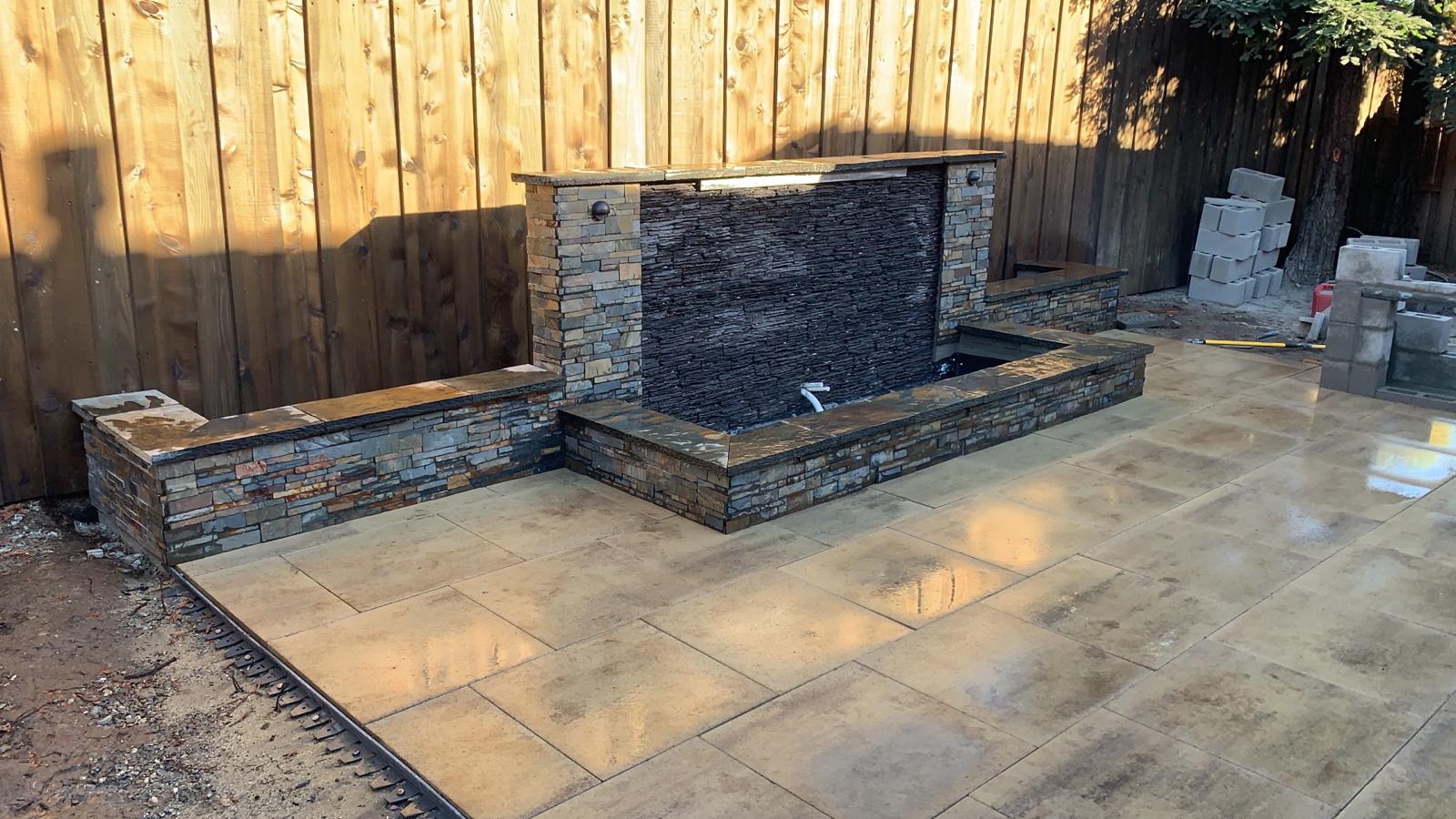 Stone water feature with dark stones, next to a wooden fence on a patio.