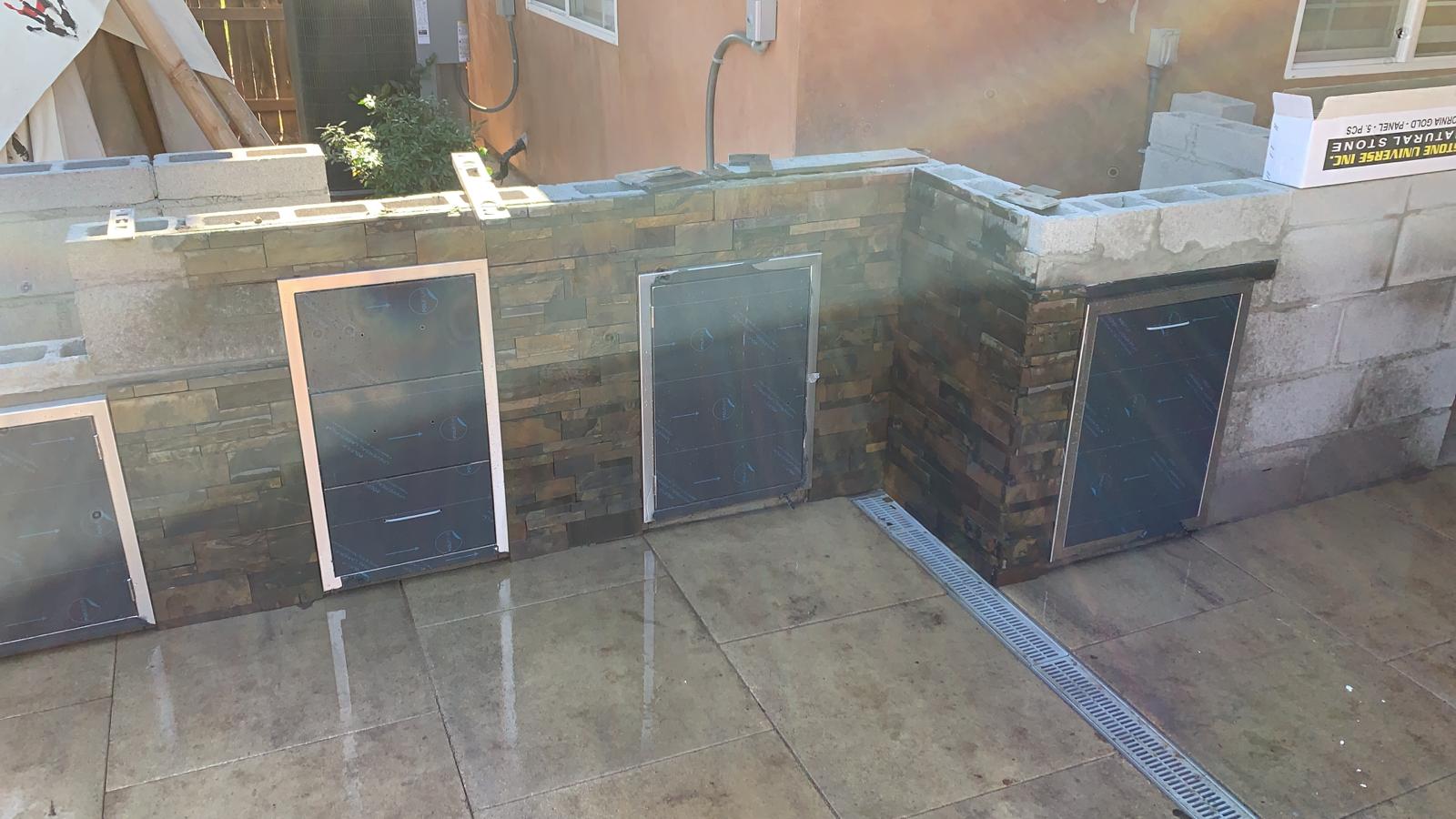 Exterior building under construction, grey block walls, dark door frames, water on the surface, sunny day.