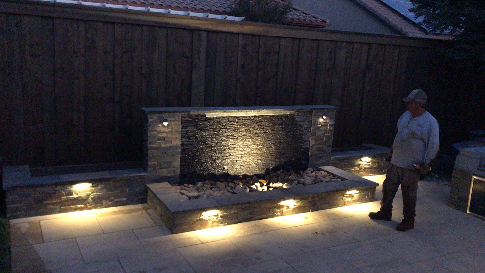 Water fountain with lighting and a person standing next to it on a patio at night.