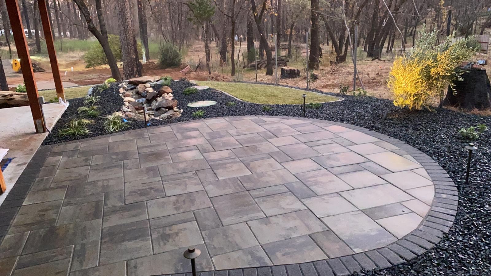 Circular paved patio with black rock border and small waterfall in a wooded setting.