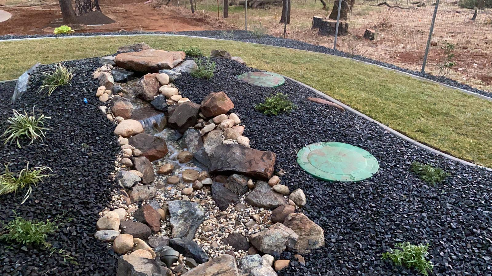 Water feature with dark stone and rocks, surrounded by black mulch, and grassy border.