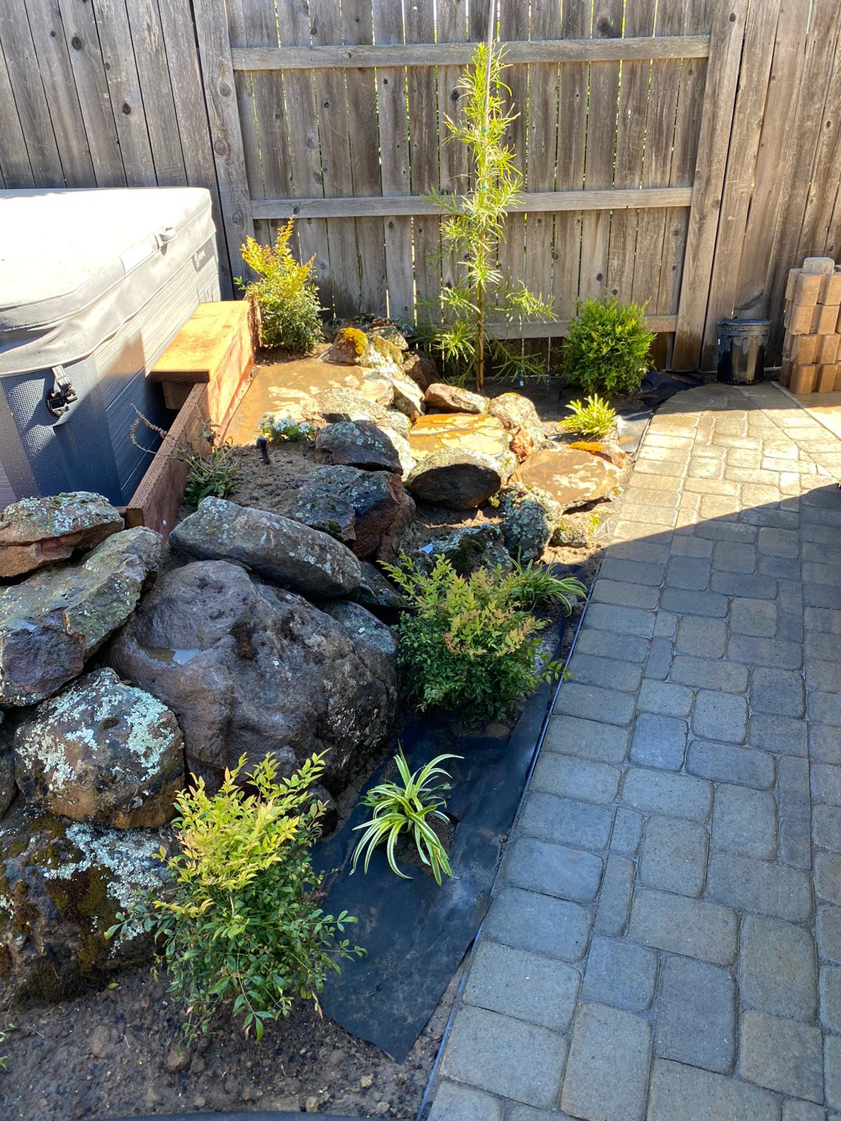 Backyard water feature with rocks, plants, and a wooden bench next to a paved patio and a wooden fence.