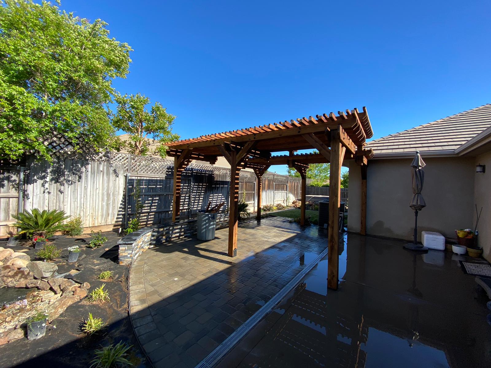Wooden pergola over a wet concrete patio in a backyard with a blue sky.