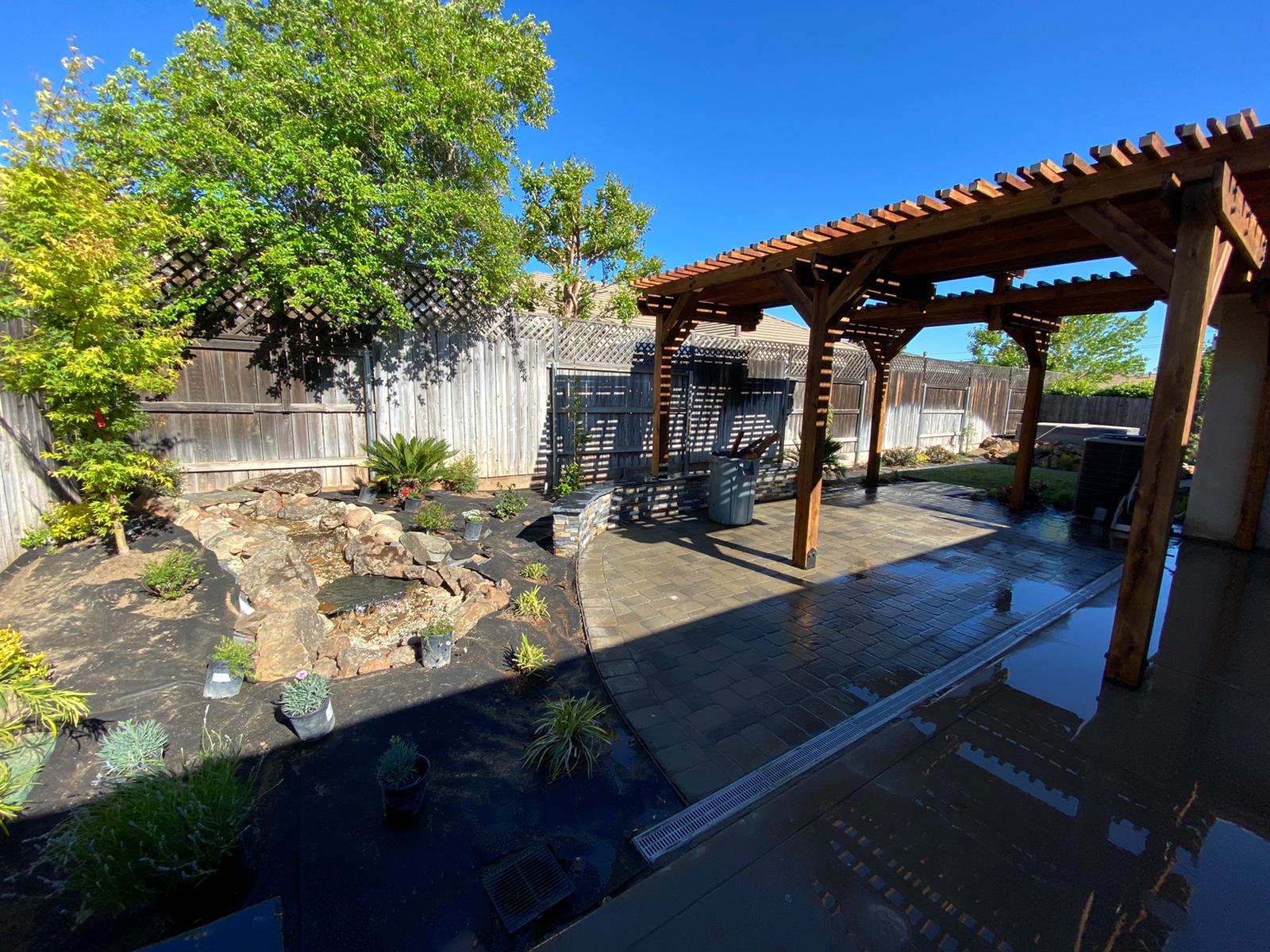 Backyard patio with wooden pergola, pond, and landscaped area under a blue sky.