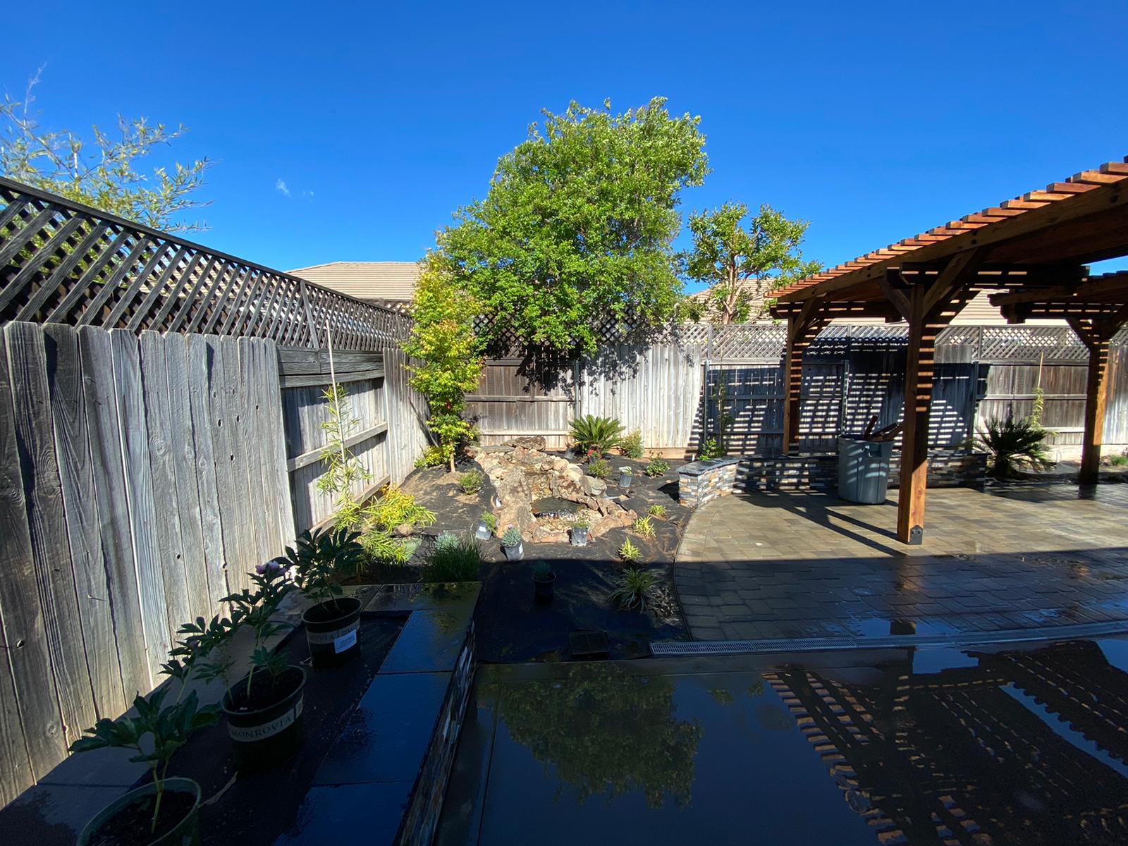 Backyard with wooden fence, patio, gazebo, and a small water feature under a bright blue sky.