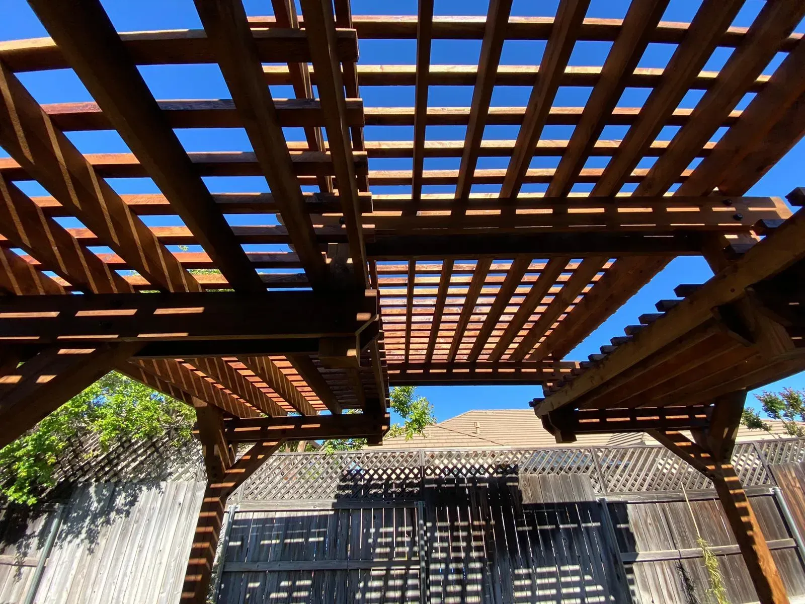 Wooden lattice pergola against a bright blue sky, casting shadows on a wooden fence.