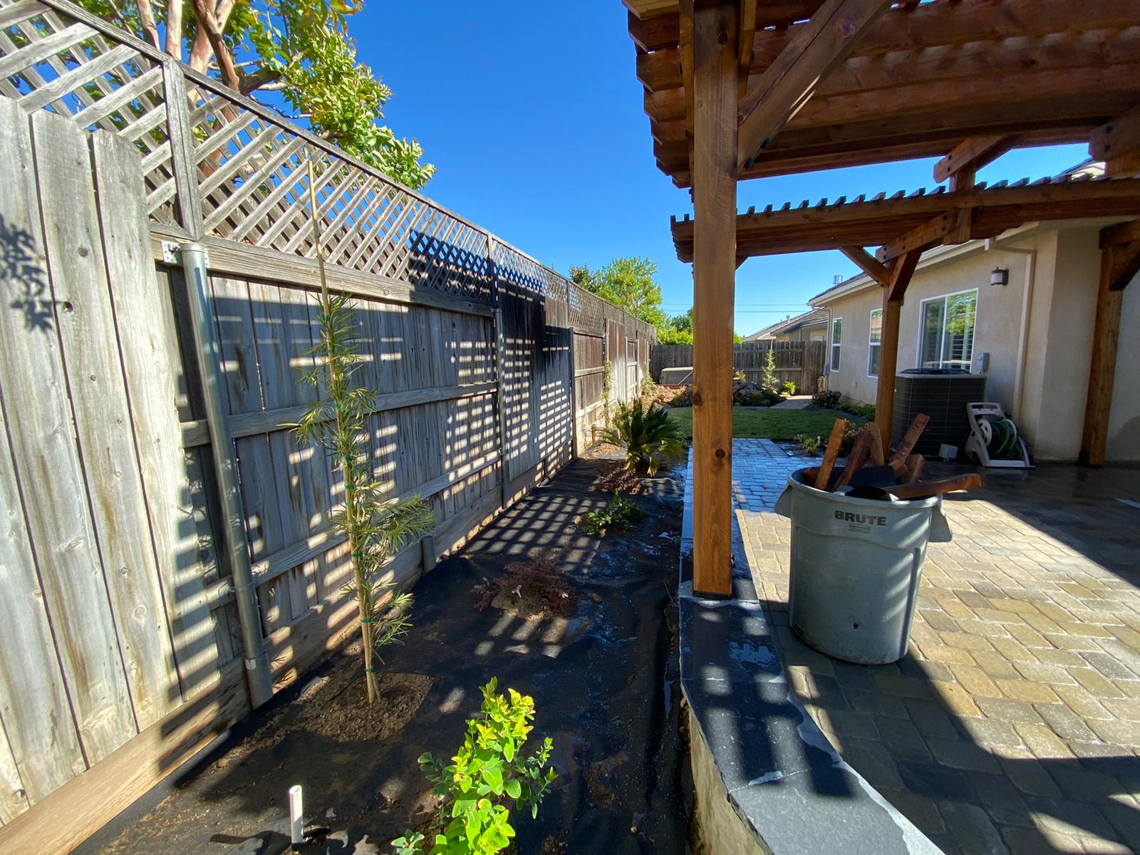 Backyard with wooden fence, pergola, and paved patio. Sunlit with shadows and green plants.