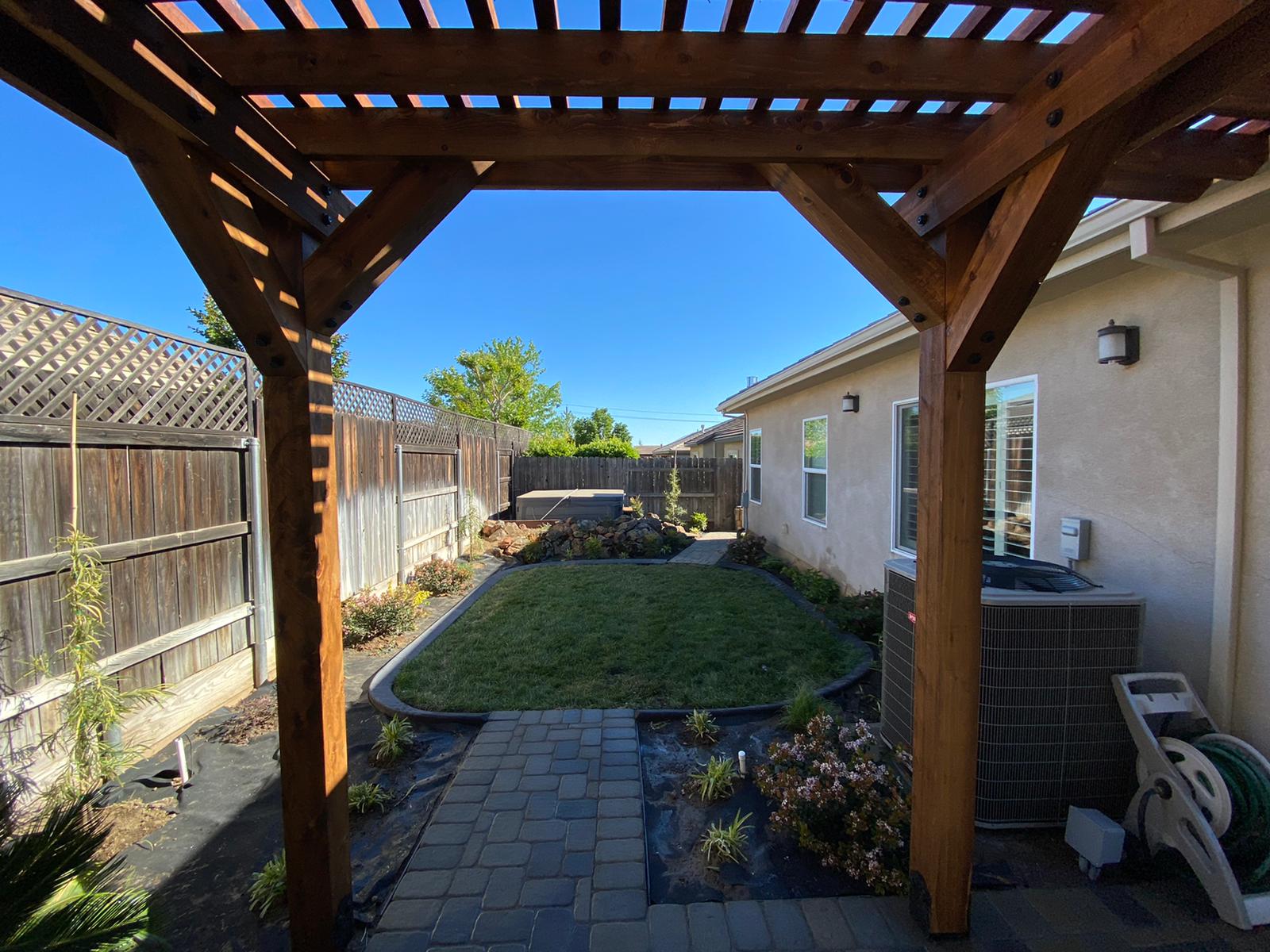 Wooden pergola frames a backyard scene: brick path, green grass, garden beds, and two-story houses under a blue sky.