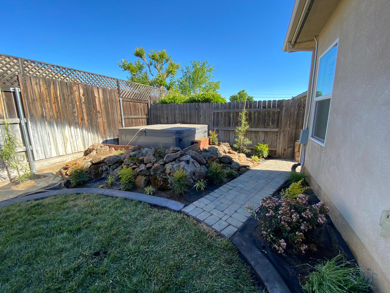 Backyard with hot tub, stone landscaping, brick walkway, wooden fence, and green grass under a blue sky.