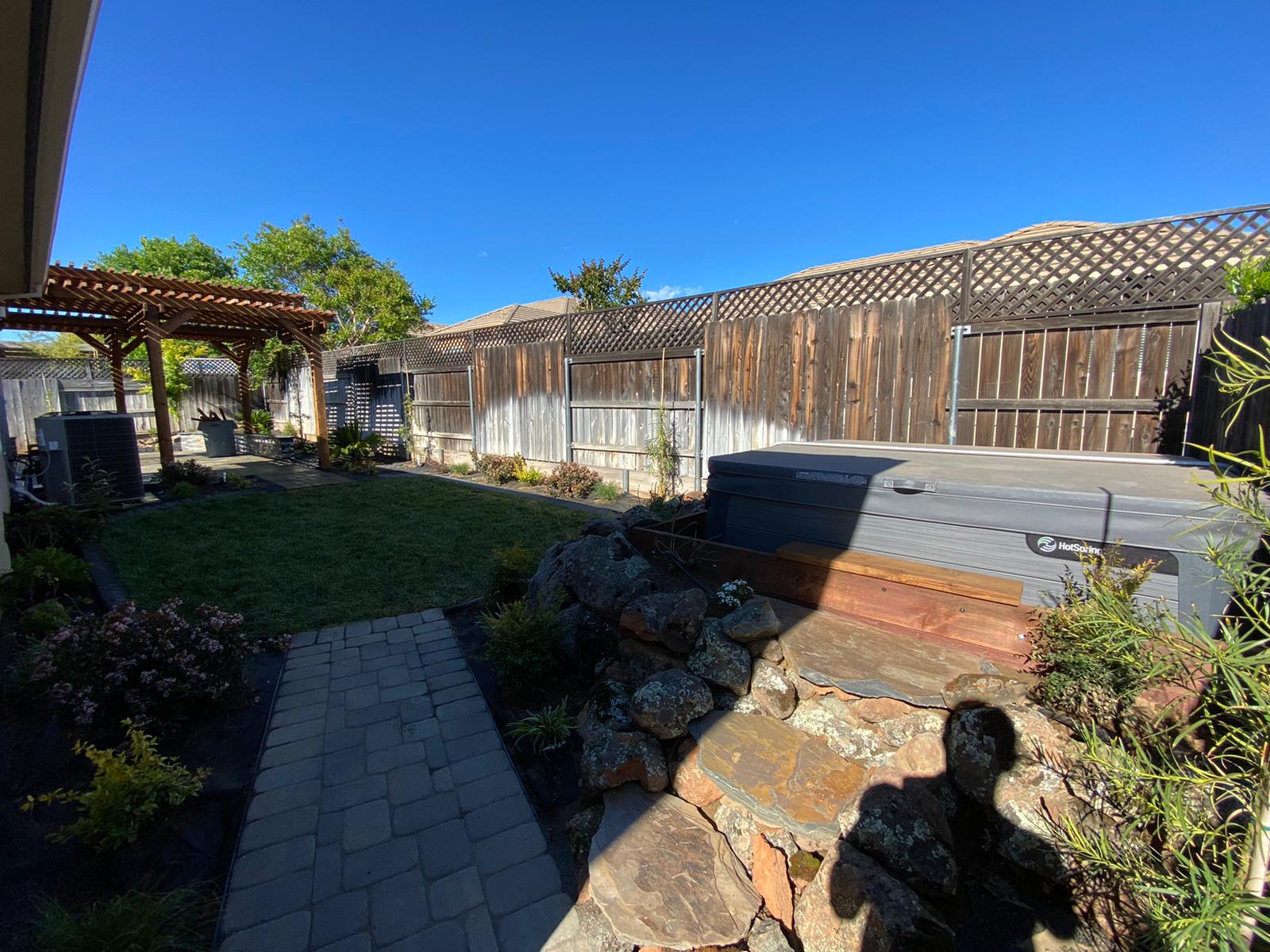 Backyard with a hot tub, stone pathway, grass, wooden fence, and pergola under a blue sky.