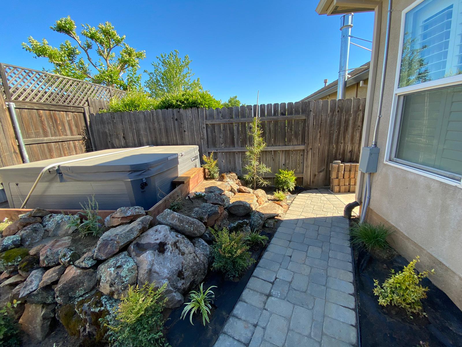 Backyard with a hot tub, rock garden, paved walkway, and wooden fence on a sunny day.