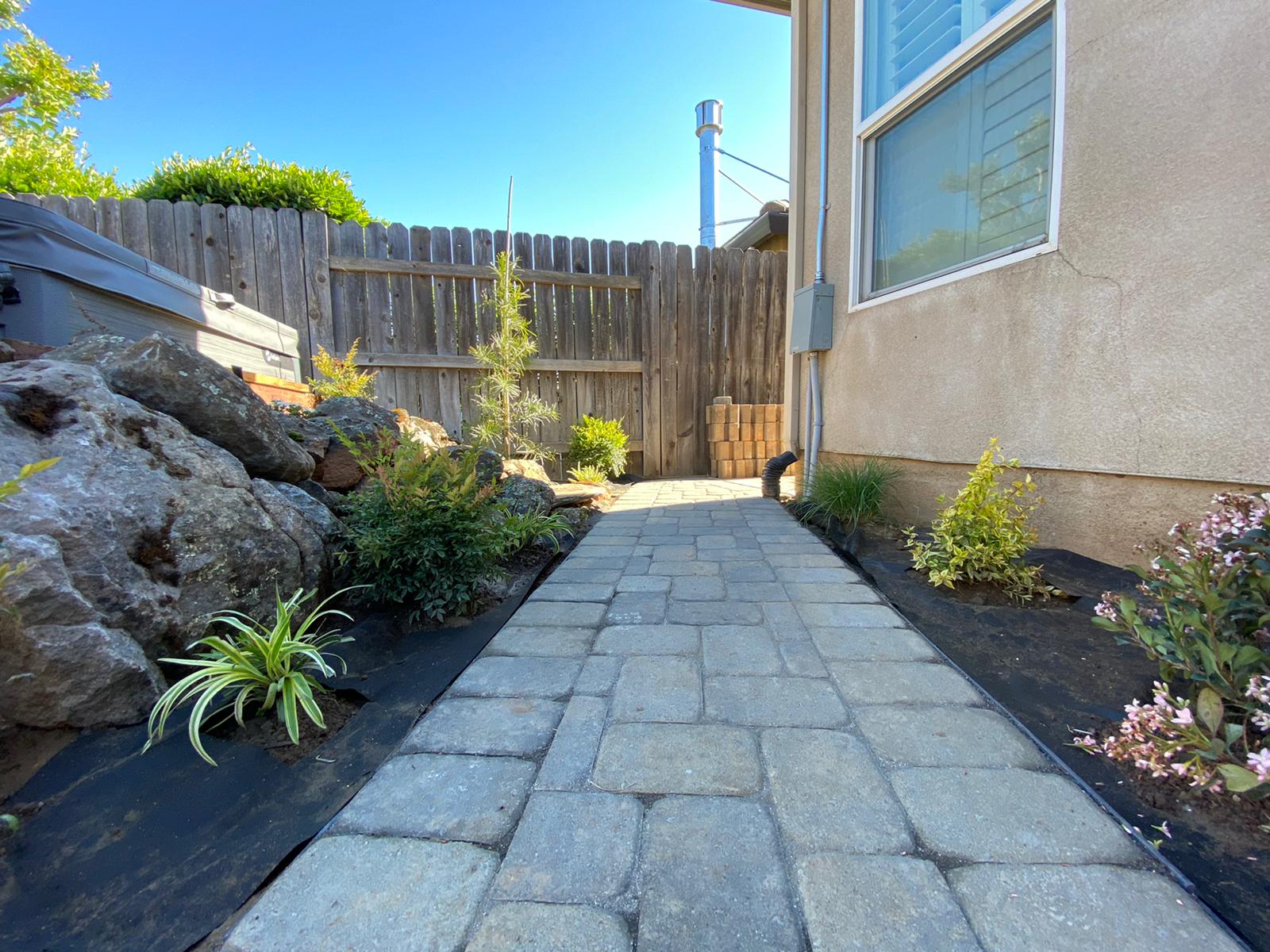 Stone paver walkway flanked by garden beds, leading to a wooden fence. House on right side.