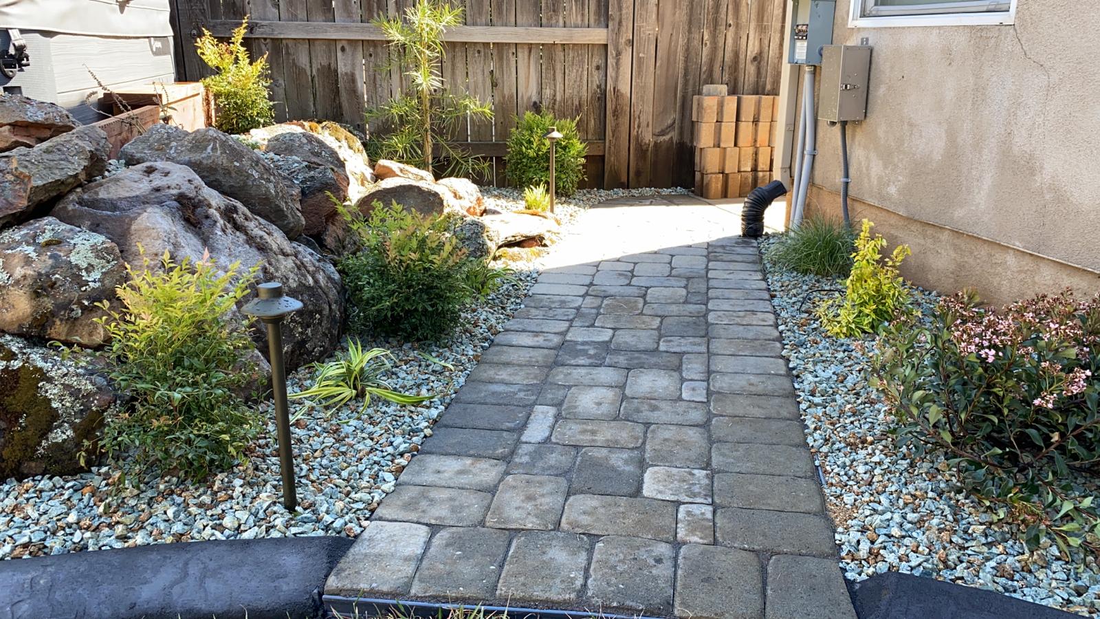 Stone pathway winds through a landscaped backyard with rocks, plants, and a wooden fence.