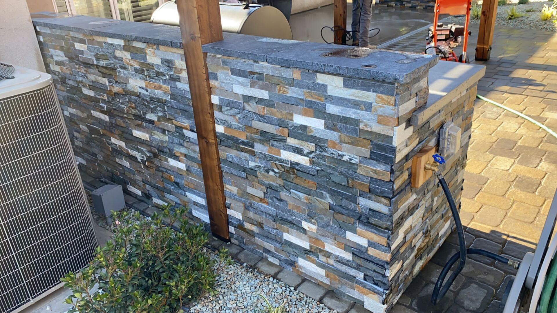 Stone-clad outdoor wall with a gray countertop and a hose. A/C unit is visible on the left.