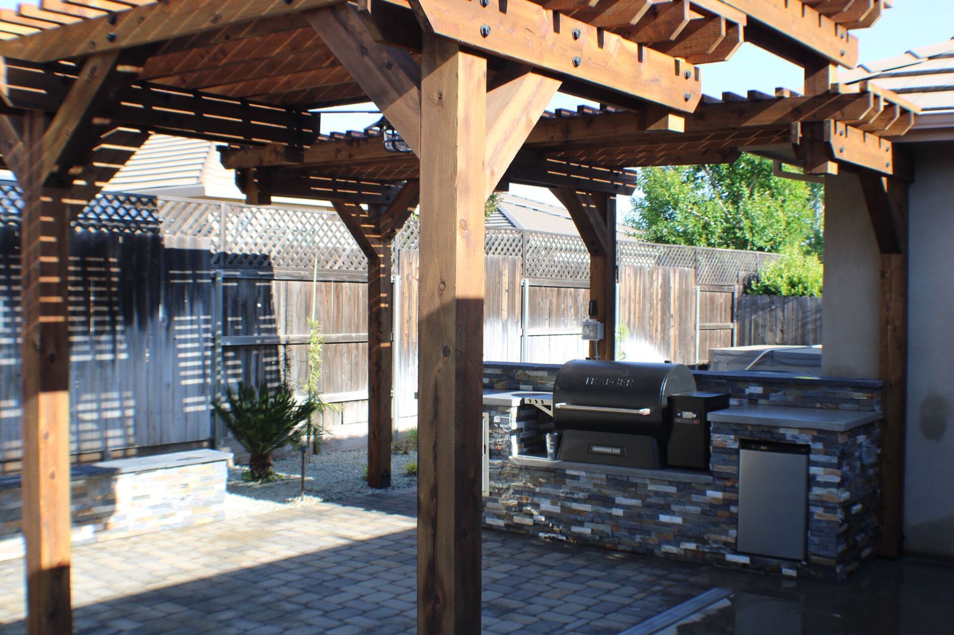 Wooden pergola over an outdoor kitchen with a grill and stone accents.
