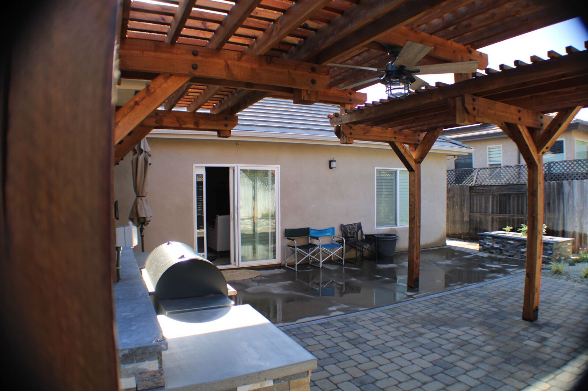 Patio with wooden pergola, brick patio, and back of a house with sliding glass door.
