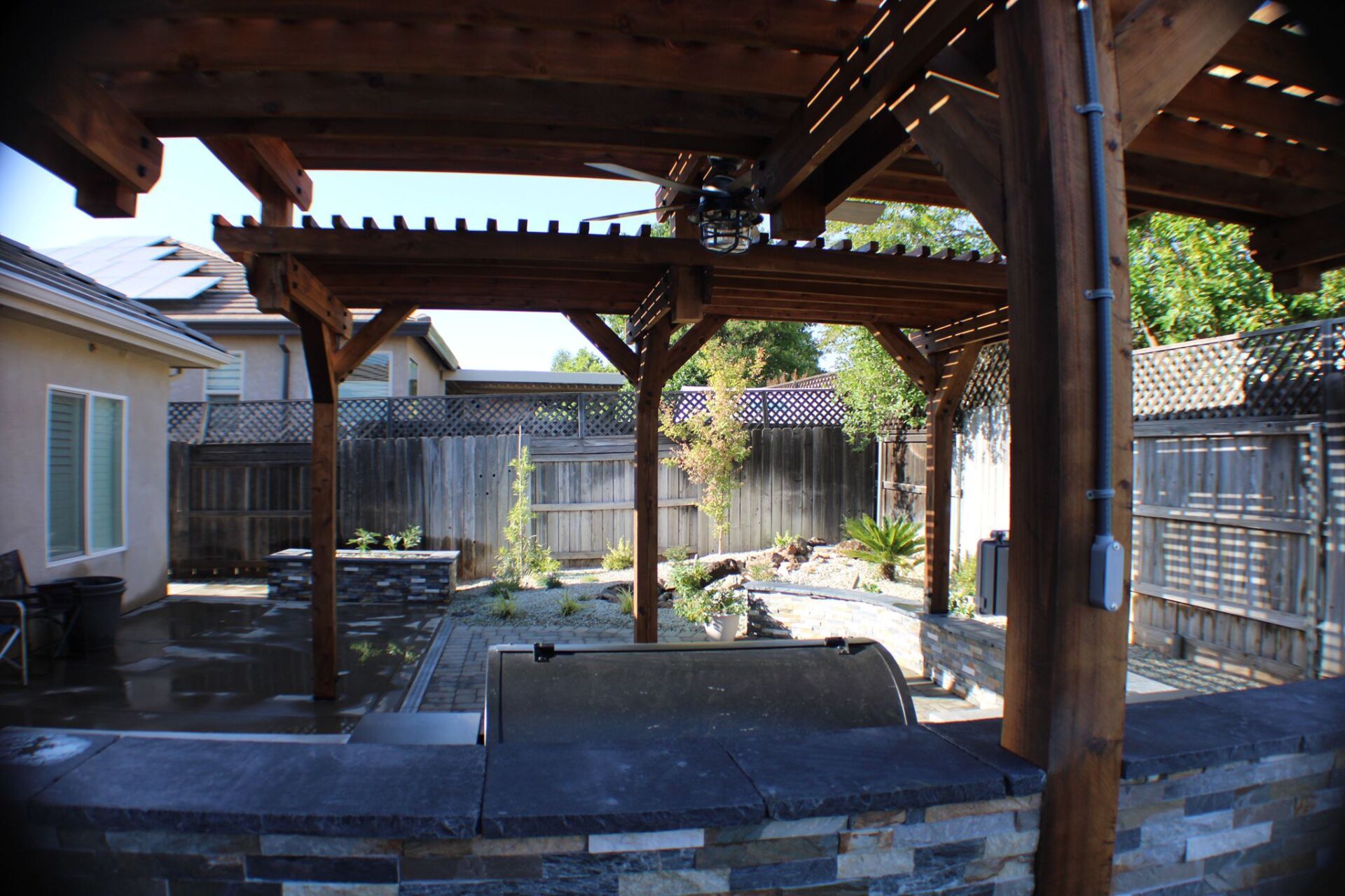 Wooden pergola in backyard with built-in grill and stone counter; fenced yard in background.
