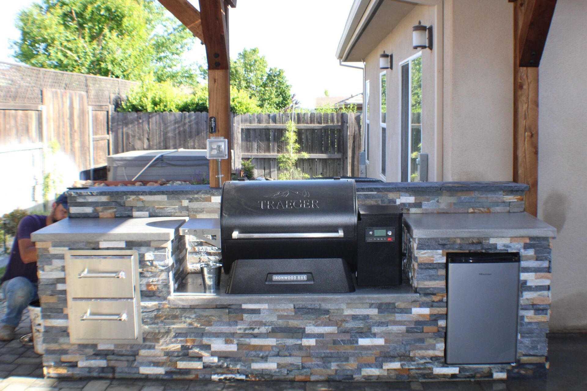 Outdoor kitchen with a built-in grill, stone facade, and a person working on the left.