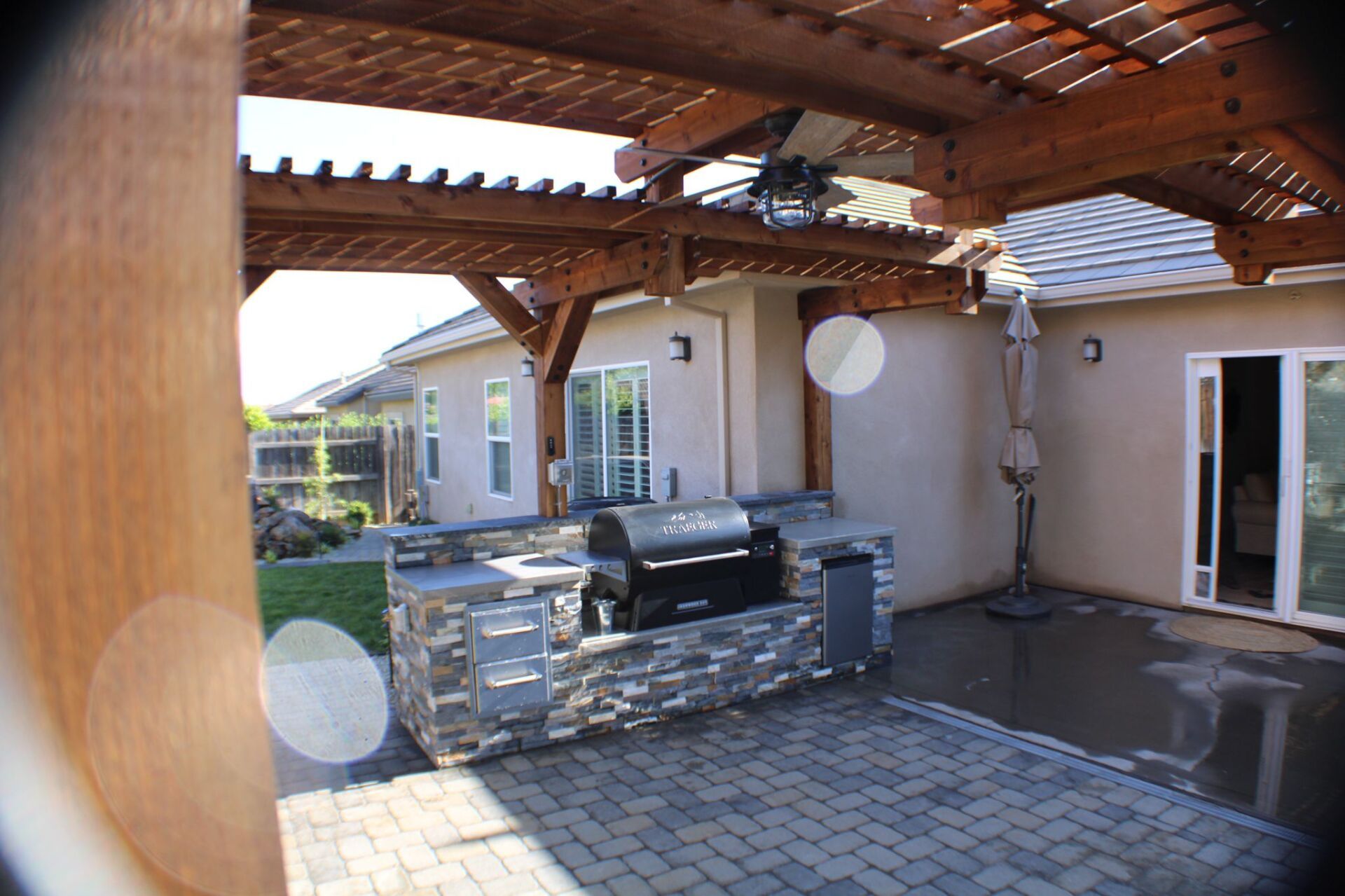 Backyard patio with built-in grill, stone facade, wooden pergola, and brick paving.