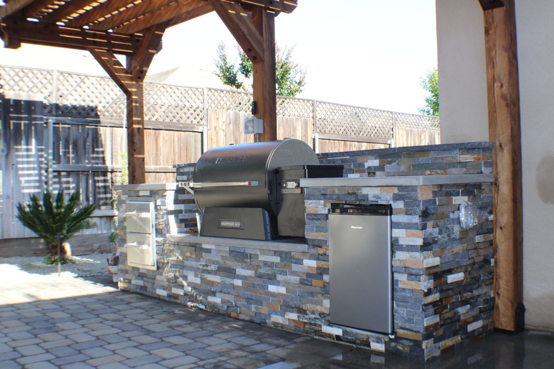 Outdoor kitchen with a built-in grill, refrigerator, and stone veneer. Underneath a pergola structure.