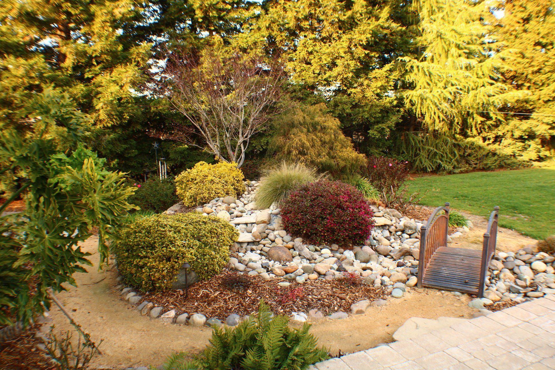 Colorful garden bed with rocks, bushes, small bridge, and lush trees.