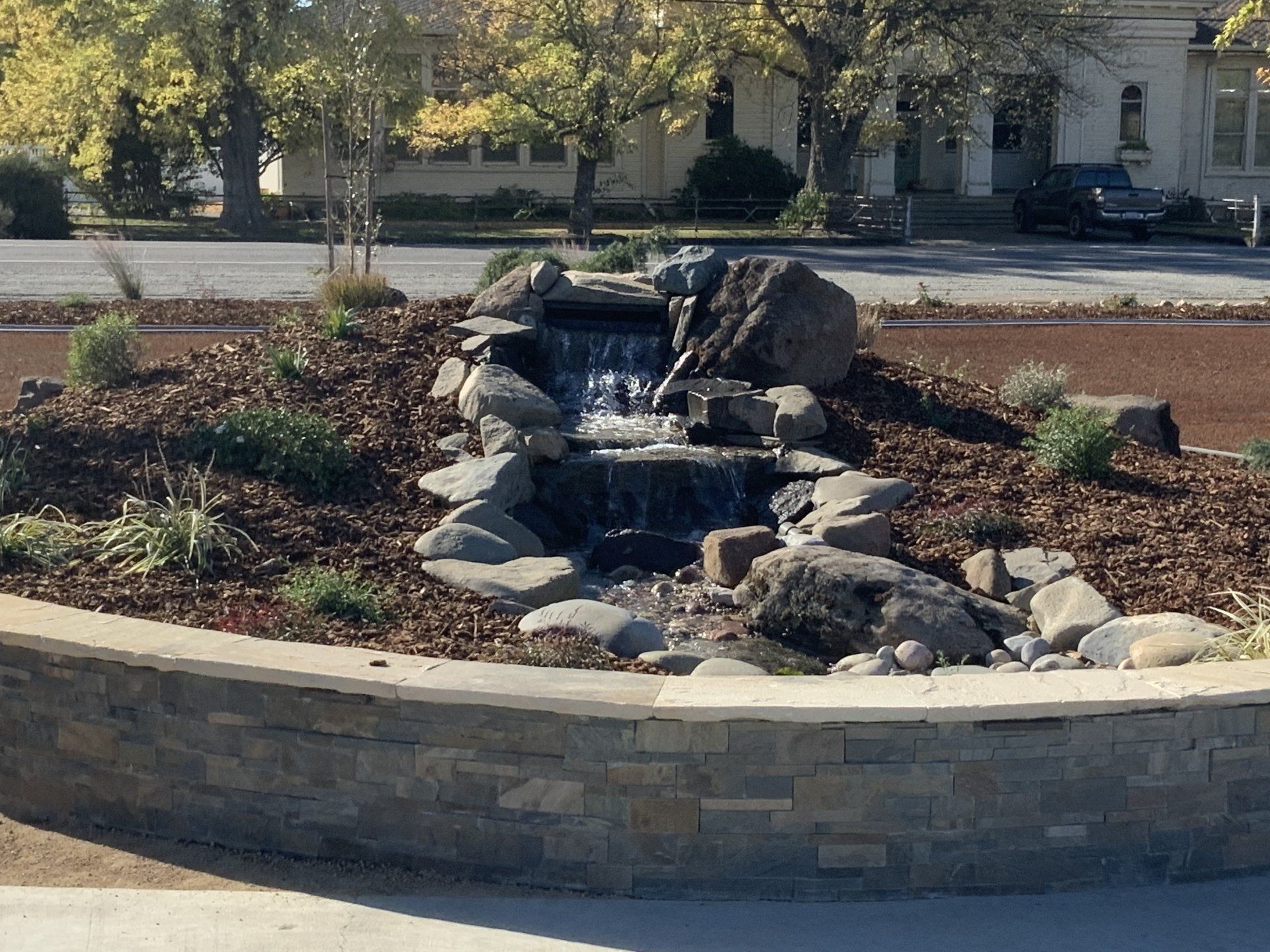 Small waterfall feature in a circular garden bed with stone wall, trees, and street in the background.