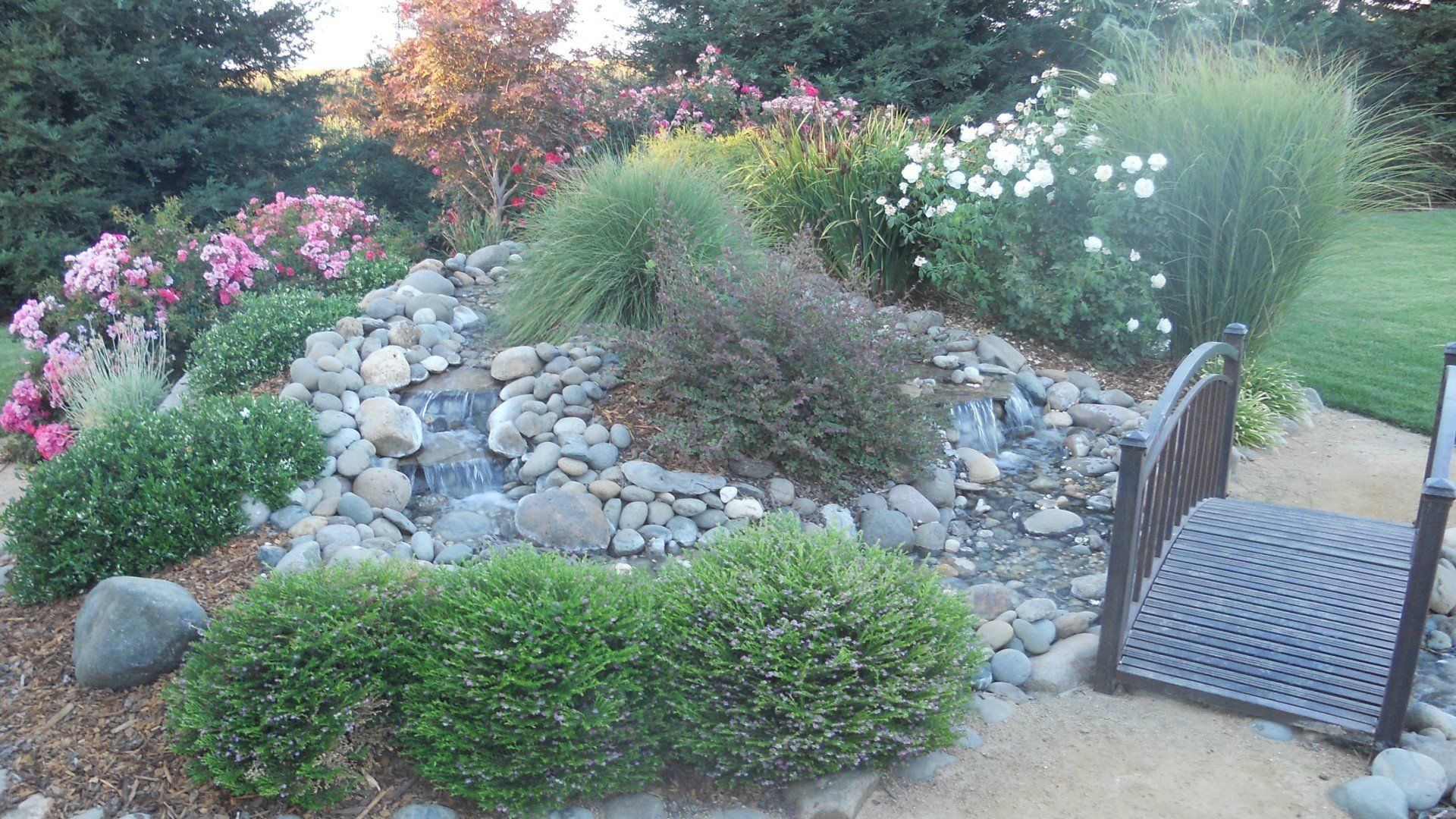 Small waterfall feature in garden bed with plants, rocks, and an adjacent chair.