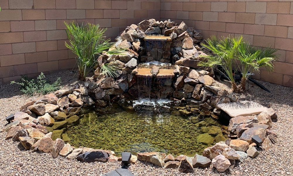 Water feature with a small waterfall surrounded by rocks and plants in a backyard setting.