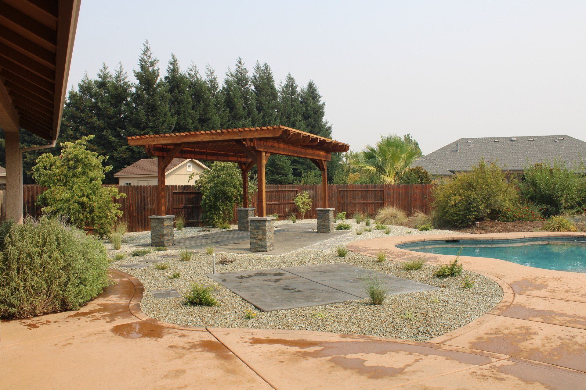 Backyard with a wooden pergola, pool, landscaping, and a cloudy sky.