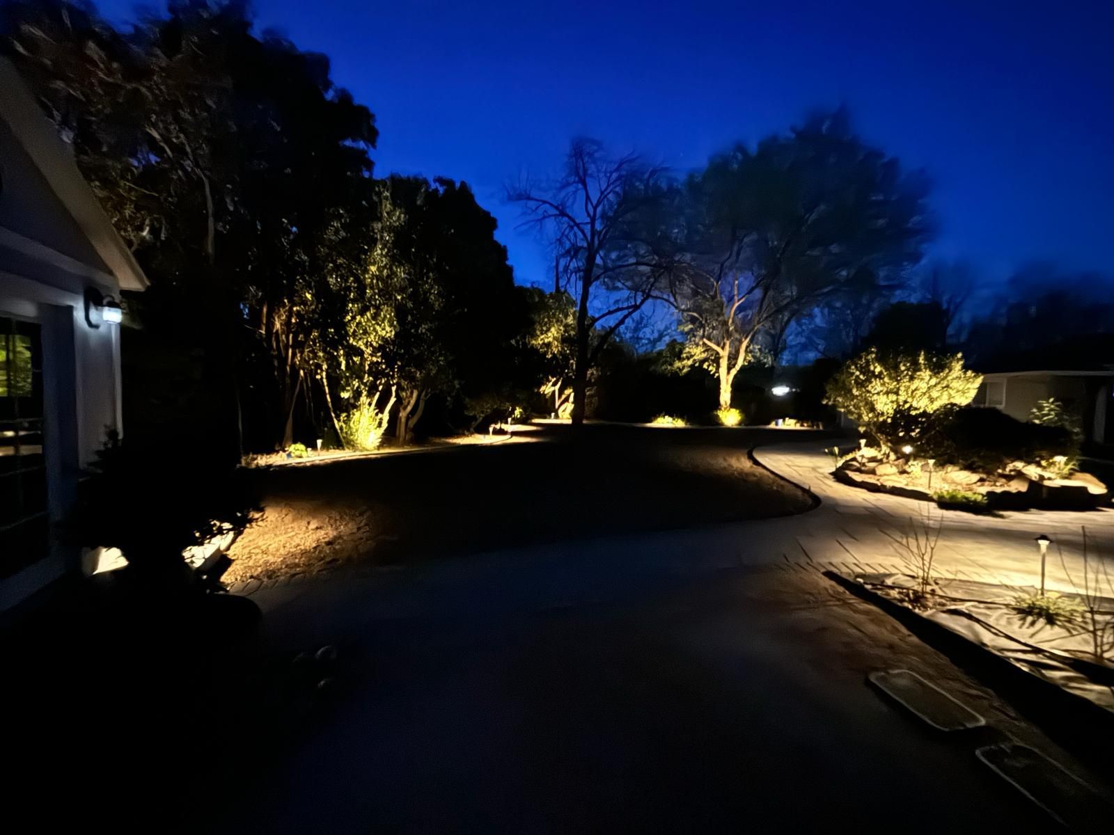 Exterior nighttime view of a house and yard illuminated by landscape lighting. Dark blue sky overhead.