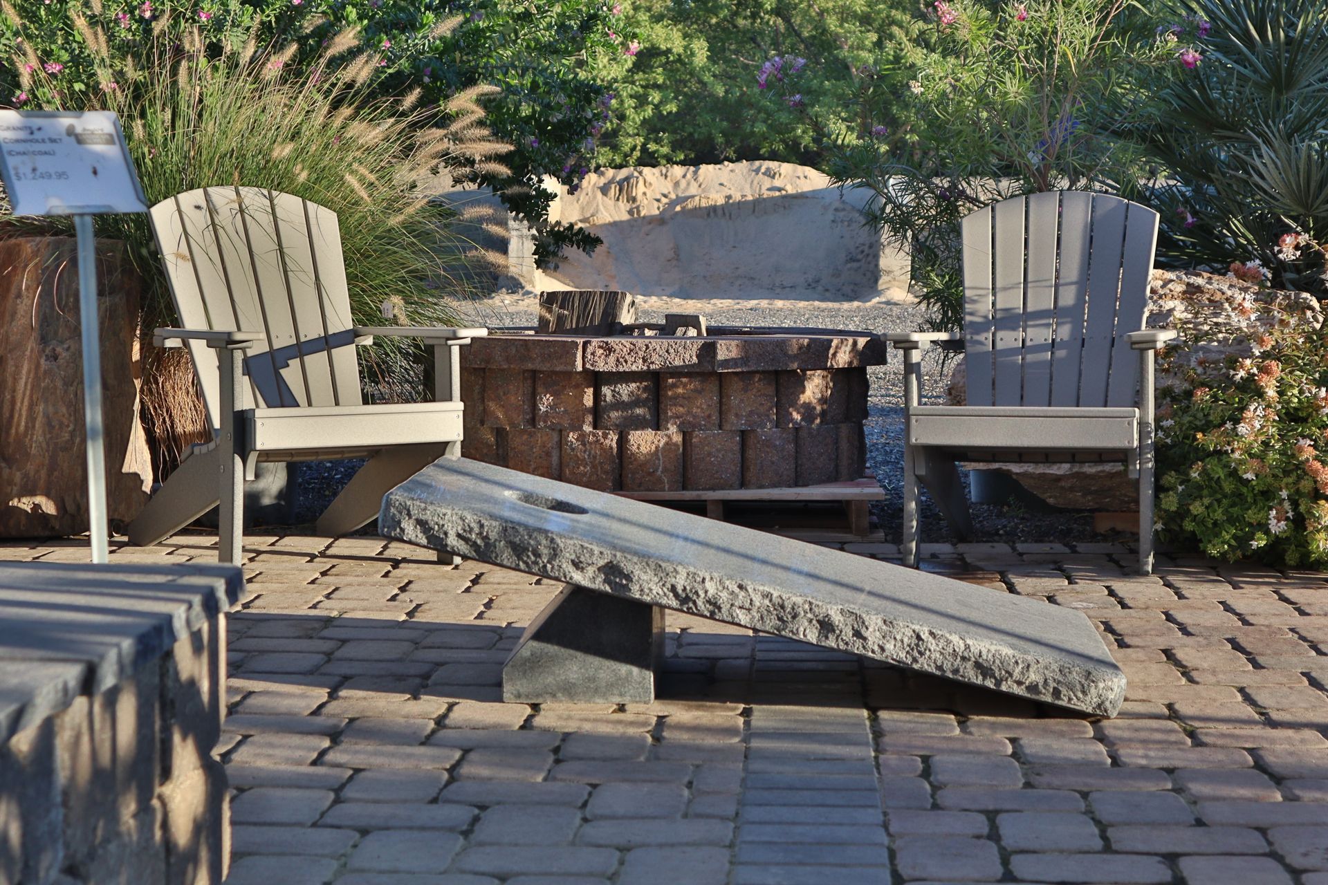 Two gray Adirondack chairs flank a stone fire pit. A stone seesaw rests on brick patio.