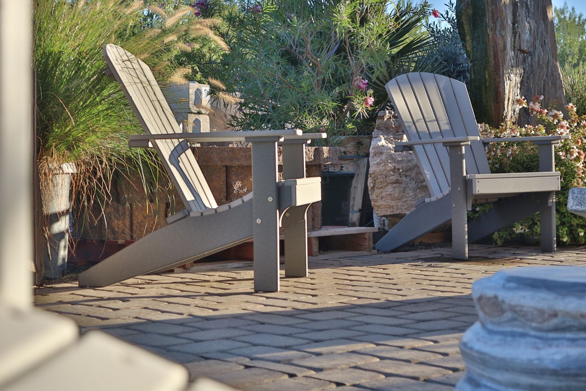 Two gray Adirondack chairs on a brick patio, set against a backdrop of greenery and a stone wall.