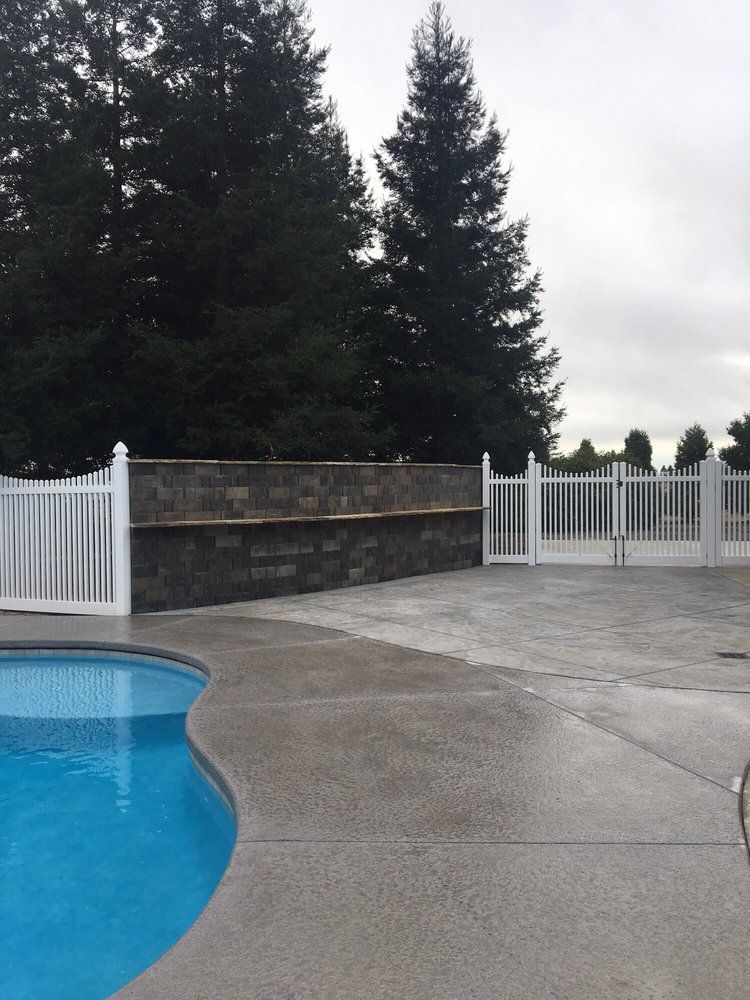 Poolside view with pool, gray concrete patio, white fence, and tall trees.