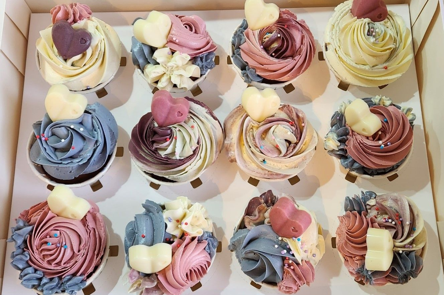 Box of twelve decorated cupcakes with pastel frosting swirls and heart-shaped candies.