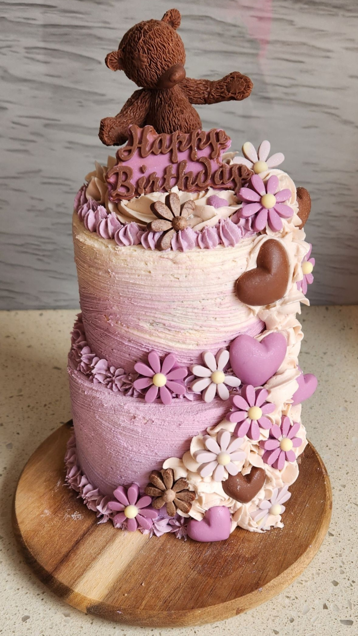 Purple and white tiered birthday cake decorated with flowers, hearts, and a teddy bear on a wooden base.