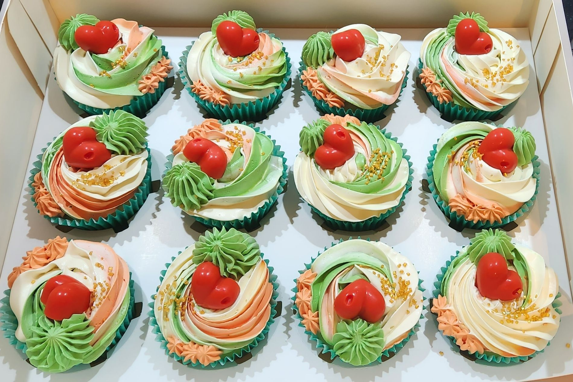 Box of frosted cupcakes with red heart decorations, various pastel colors.
