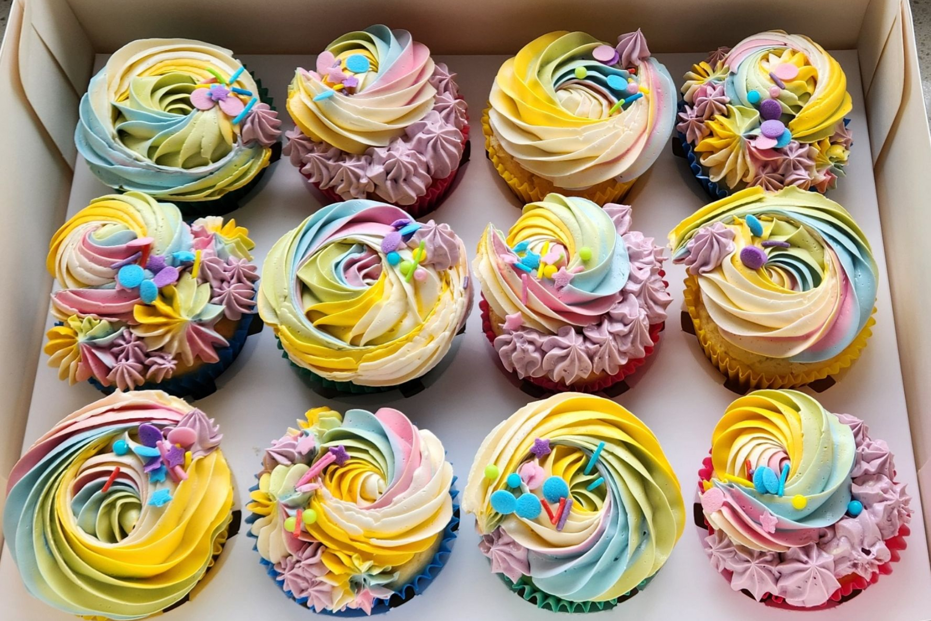Box of colorful cupcakes with swirled frosting in shades of yellow, blue, and pink, decorated with sprinkles.