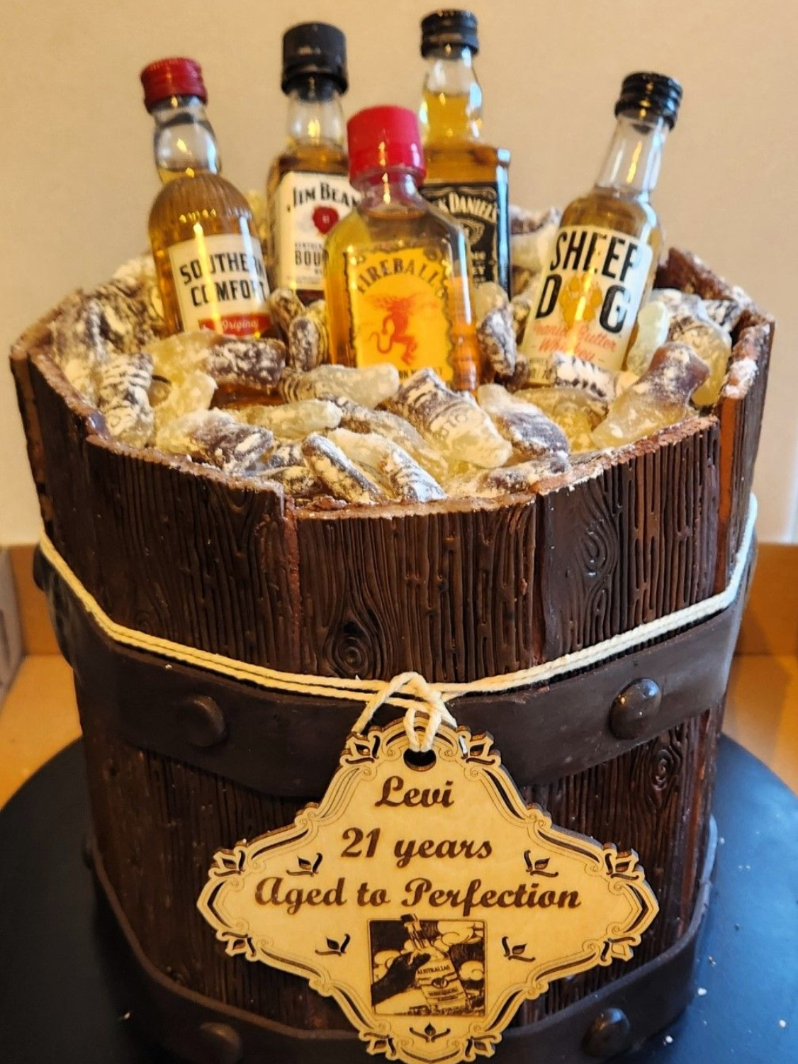 Cake in the shape of a wooden barrel with liquor bottles and a personalized tag that says Levi, 21 years old.