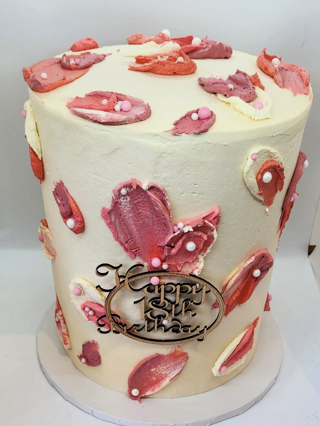 Tall white frosted cake decorated with pink, red, and white abstract shapes, “Happy Birthday” plaque.