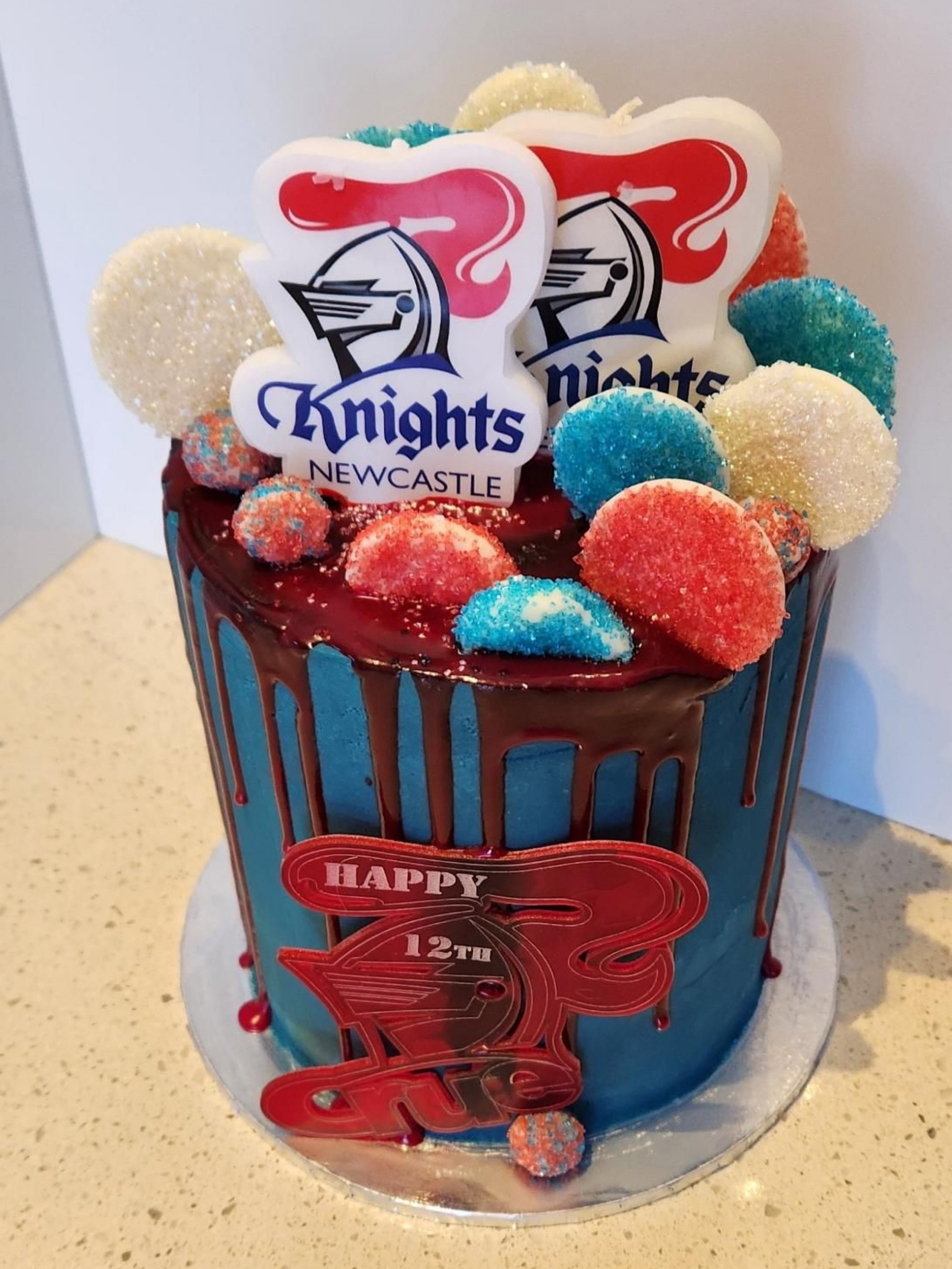 Cake decorated with a blue icing, dripping chocolate, and Newcastle Knights logos and candies. 