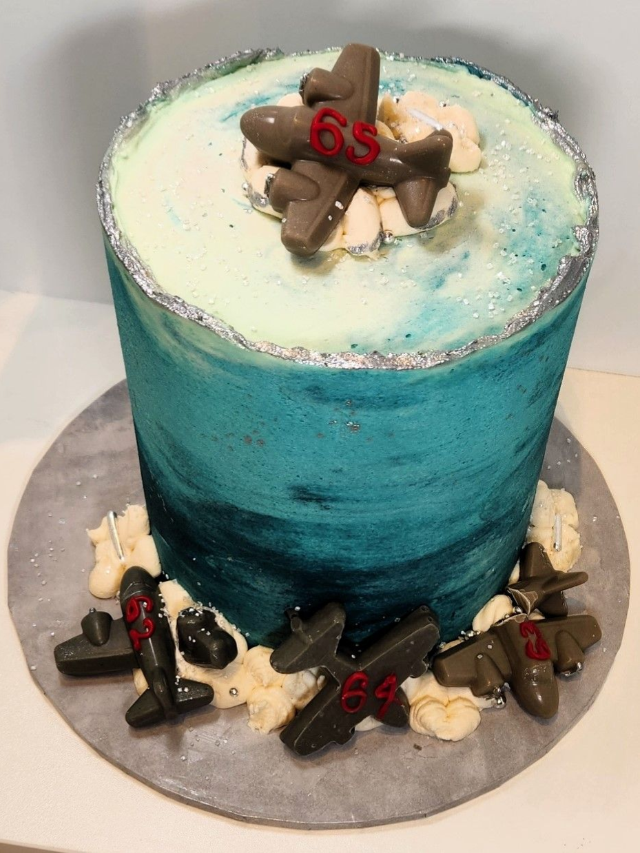 Cake decorated with blue-green frosting and chocolate airplane accents, celebrating 65 years. Silver glitter trim.