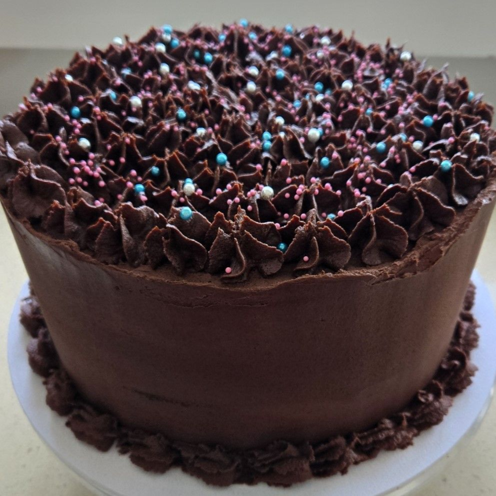 Chocolate cake with chocolate frosting, topped with sprinkles, on a white cake stand.