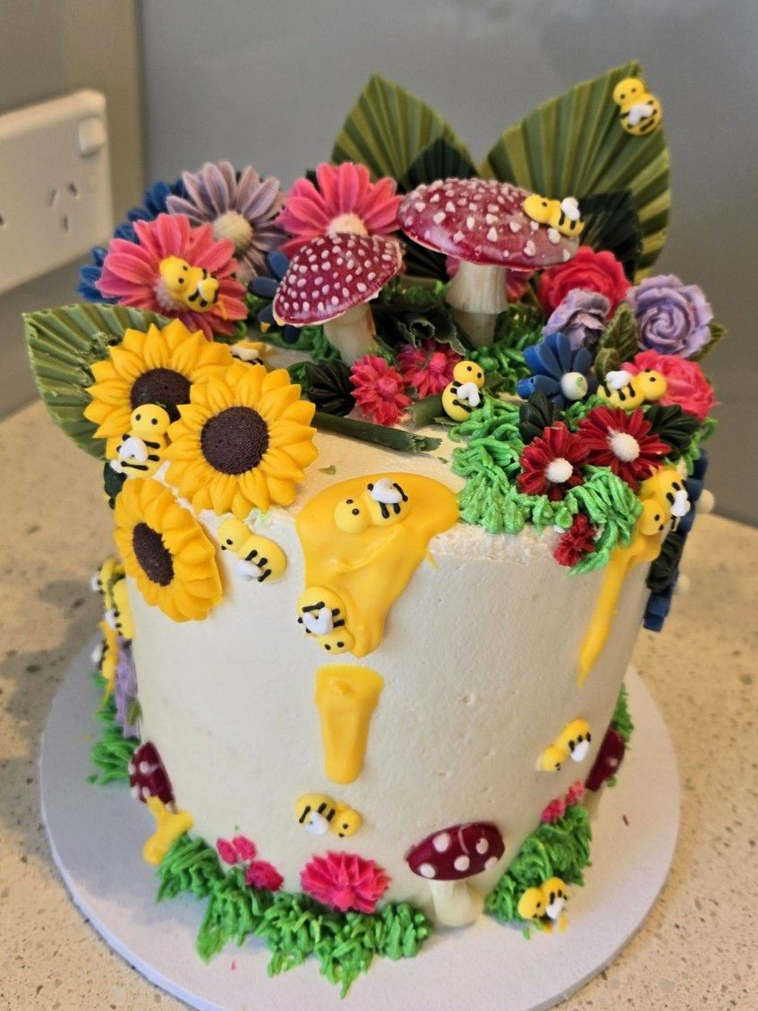 Cake decorated with flowers, mushrooms, bees, and yellow honey drips.