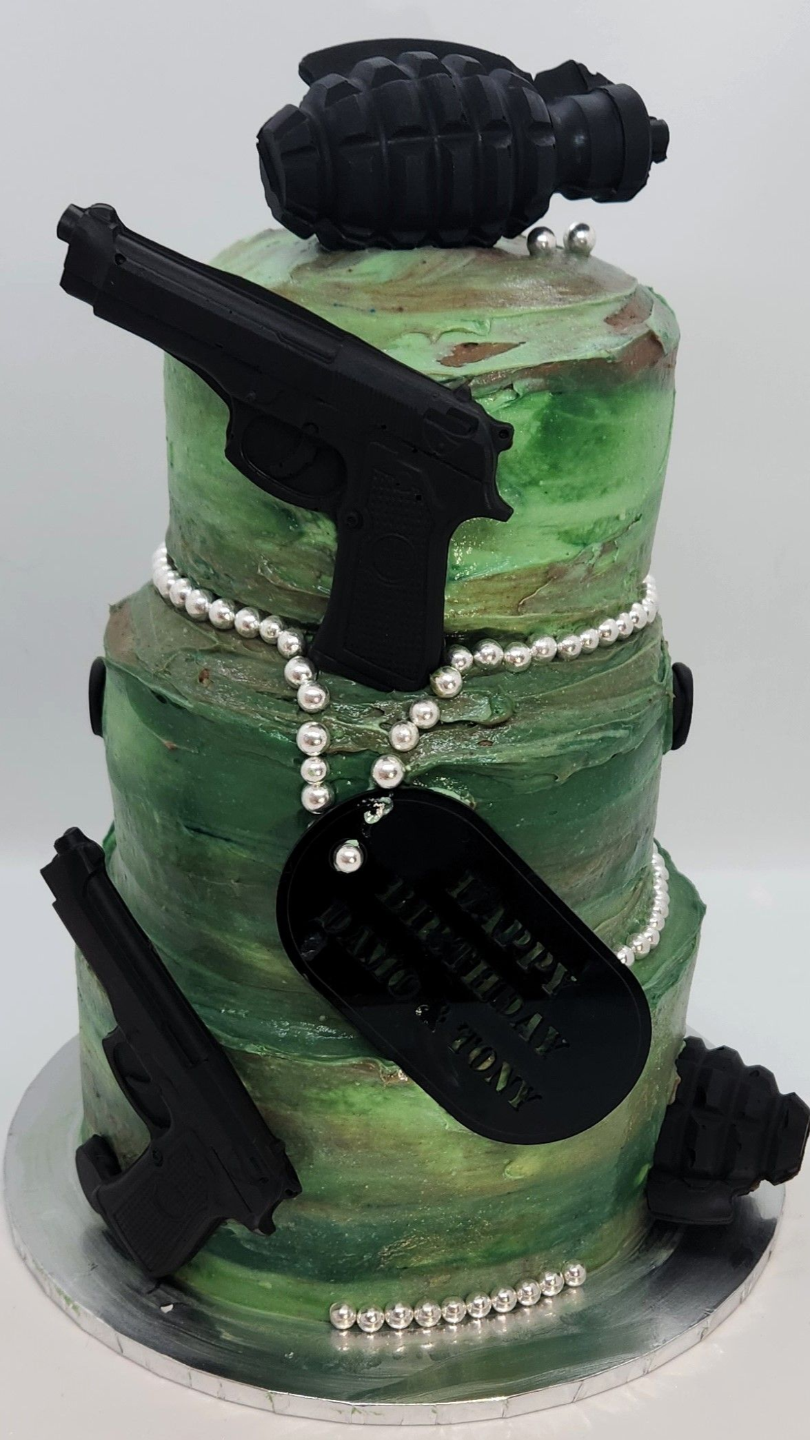 Three-tiered green cake decorated with black gun cutouts, grenades, a dog tag that reads 